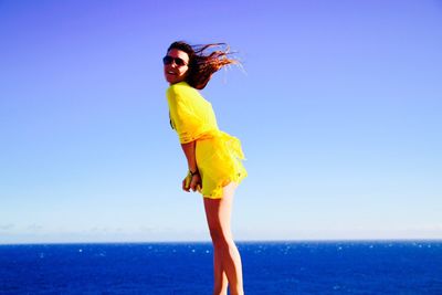 Side view of happy woman in yellow top standing at beach against sea and sky on sunny day