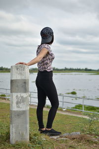 Side view of young woman standing on grassy field against cloudy sky