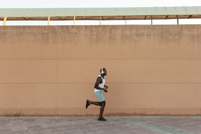 Side view full body of energetic african american sportsman in headphones jogging along pavement during fitness training