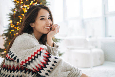 Portrait of young asian woman with dark long hair in cozy sweater in room with christmas tree