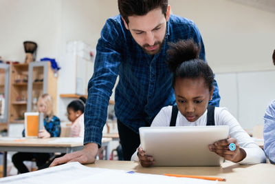 Teacher looking at girl using digital tablet in classroom