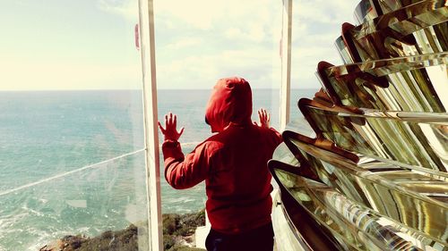 Rear view of boy in red hood looking through window at lighthouse