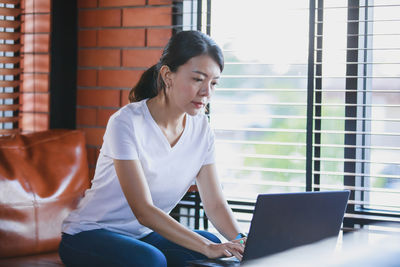Young woman using phone while sitting on window