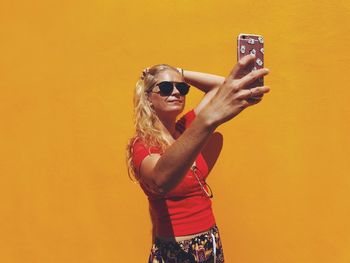 Woman taking selfie while standing against orange wall during sunny day