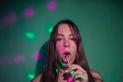 Portrait of young woman licking lollipop while lights falling on her against green background