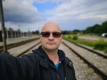 Portrait of man wearing sunglasses on railroad tracks against sky