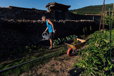 Full body side view of male farmer with watering can pouring water on vegetables while standing near wall with plants