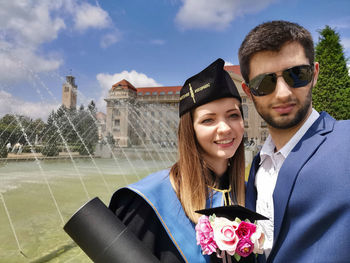 Young woman wearing graduation gown with man by fountain