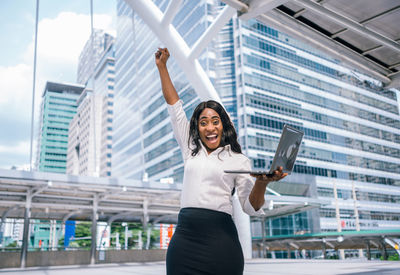 Portrait of smiling businesswoman with laptop standing against building
