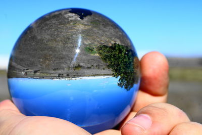 Cropped hand holding crystal ball with reflection of landscape against clear sky