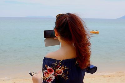 Rear view of redhead woman photographing sea against sky