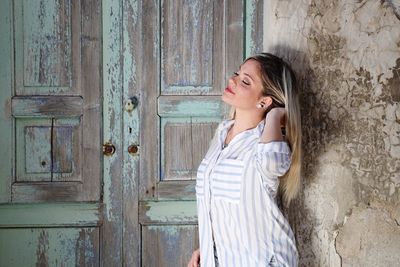 Side view of young woman with hand in hair standing against abandoned house