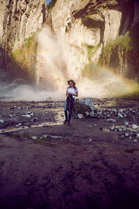 Woman in jeans and boots stands next to a waterfall in the summer in the caucasus