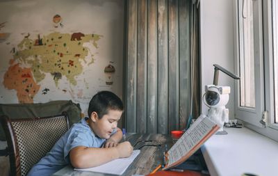 Asian boy write in notebook with geography wall map, home education, e-learning, tutoring services
