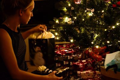 Side view of woman with gifts by illuminated christmas tree at home
