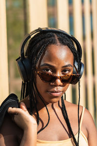 Young black woman with stylish jacket leaning on wall of contemporary building and listening to music in headphones on city street looking at camera