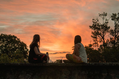 Two young women sitting by trees against sky during orange sunset in a small town