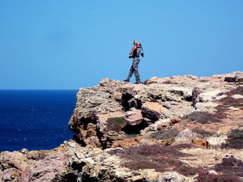 Man standing on rock by sea against clear blue sky