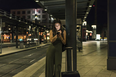 Young woman waiting at the station by night using cell phone