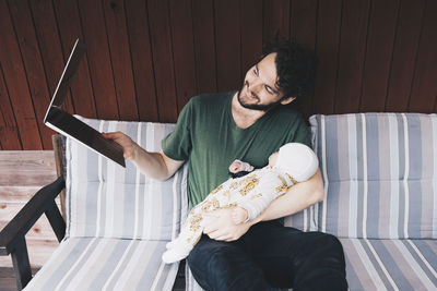 Smiling father showing baby boy in video conference on laptop while sitting at holiday villa