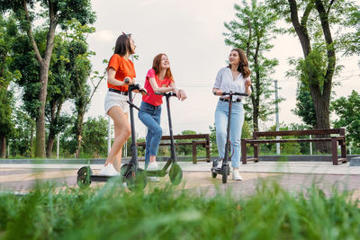 Three young girl friends on vacation having fun driving electric scooter through the city park