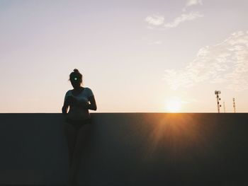 Woman using mobile phone while leaning on retaining wall against sky during sunset