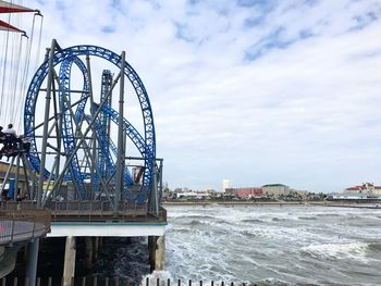 Roller coaster by sea in city against cloudy sky