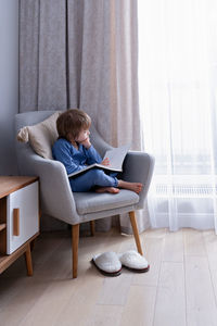 Cute preschool blonde kid reading book sitting on a cozy chair in light nordic style living room