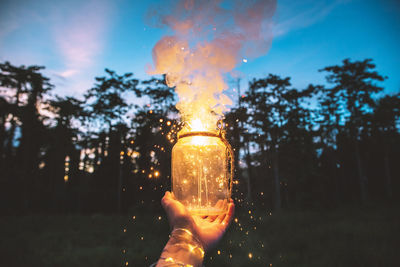 Cropped hand holding jar of lit sparklers against sky