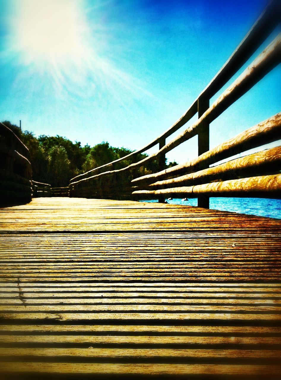 sunlight, blue, water, sun, built structure, clear sky, sky, sunbeam, railing, wood - material, pier, sunny, lens flare, surface level, nature, tree, tranquility, outdoors, boardwalk, no people