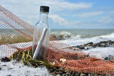 Close-up of message in bottle by fishing net at beach against sky