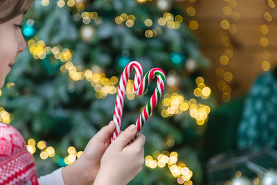 Cropped hand of girl holding candy cane against christmas tree at home