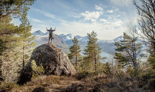 Man standing on rock formation amidst trees against cloudy sky