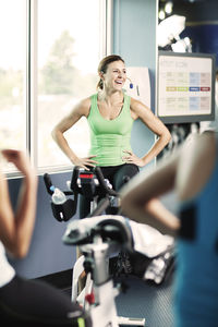 Happy trainer with hands on hip sitting on exercise bike at gym