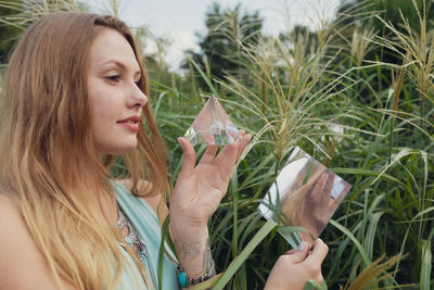 Young woman holding prism and mirror while standing against plants