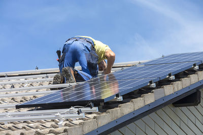 Low angle view of man installing solar panels on roof against sky