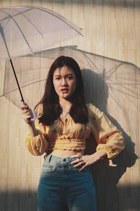 Portrait of young woman holding umbrella while standing against wall