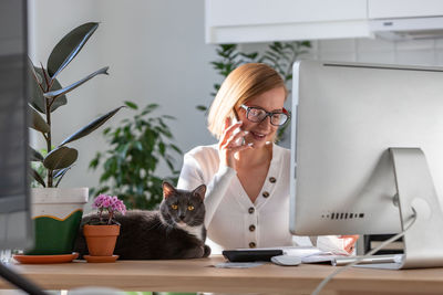 Woman talking on phone remote working on computer from home office, cat sitting on the table. 