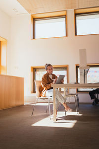 Pensive young female student with stylus leaning on hand and browsing data on tablet while sitting at table doing homework in university library