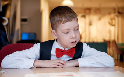 A boy in a white shirt and a red bow tie sits in a cafe and reads the menu. 