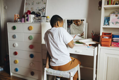 Boy writing in book while sitting on seat by table at home