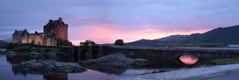 Panoramic shot old arch bridge over river by castle against sky during sunset