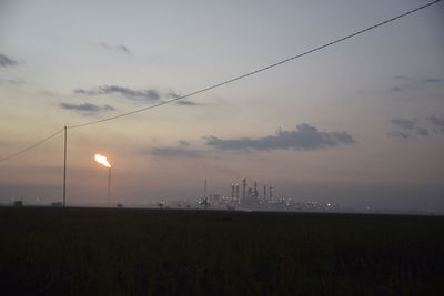 Burning flare stack and industrial buildings against sky during sunset