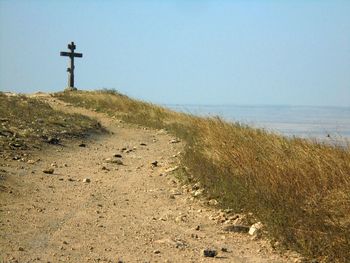 Dirt trails and cross on hill by sea against clear sky