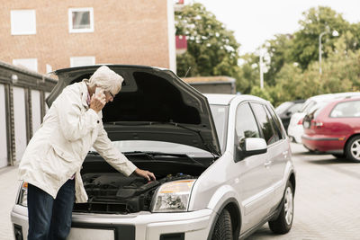 Senior woman examining car engine while using phone at roadside