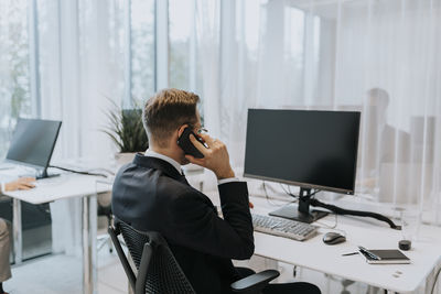 Rear view of businessman talking on mobile phone while sitting at computer desk in office