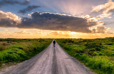 Man standing on road passing through landscape against cloudy sky