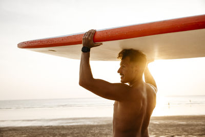 Man carrying paddleboard on head by sea at beach during sunset