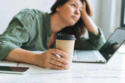Young unhappy woman plus size working at laptop on table with paper cup of coffee in office