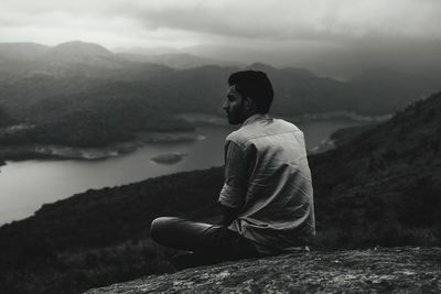 Man sitting on rock looking at mountains against sky